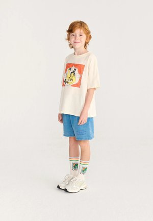 Enfant roux portant un t-shirt graphique crème, un short bleu, des chaussettes colorées avec un motif étoile et des baskets blanches, debout sur un fond uni.