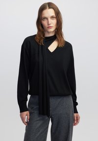 Zwarte gebreide trui met een V-hals en een asymmetrisch ontwerp, voorzien van een gedrapeerde sjaaldetail. Lange mouwen en een relaxte pasvorm.