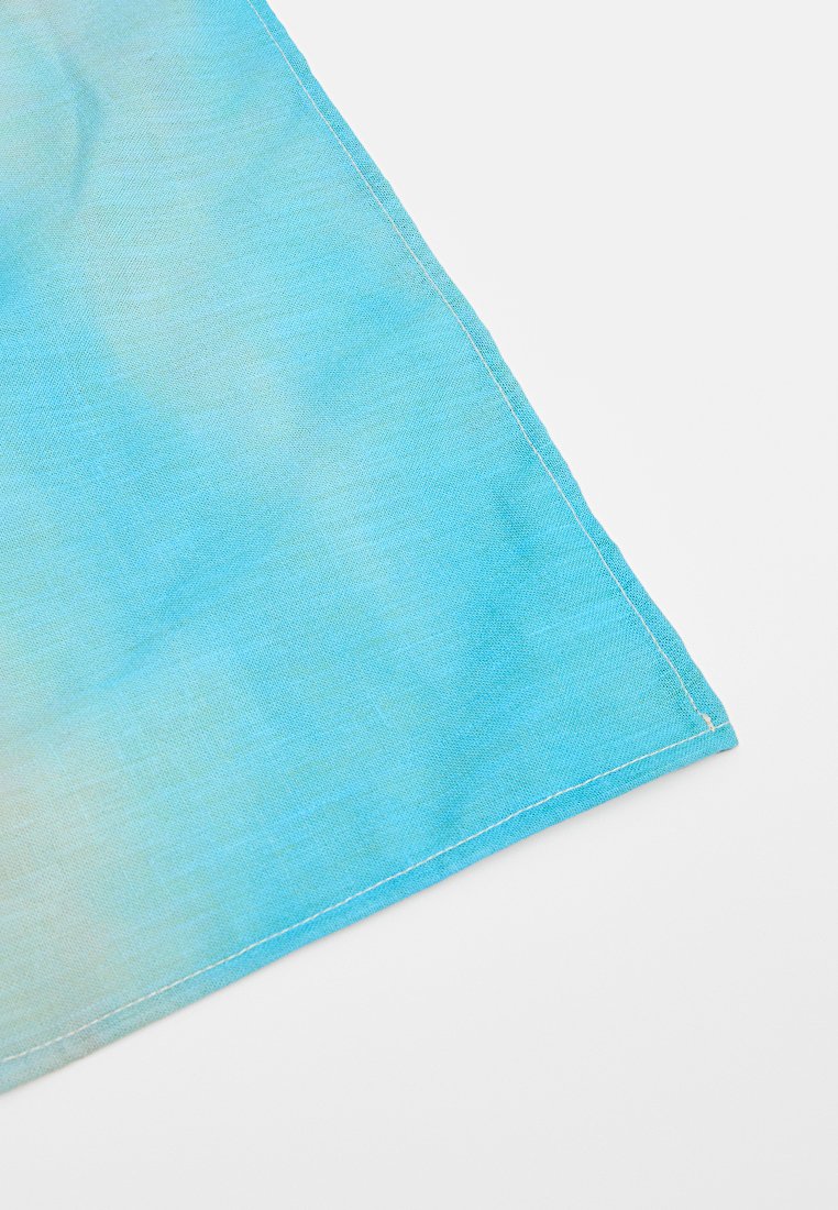 Róg niebieskiej i zielonej tkaniny gradientowej z białą haftowaną lamówką na białym tle, ukazujący teksturę i detale przeszyć.