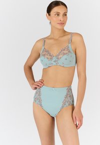 Vrouw in een lichtblauwe geborduurde beha en hoge taille slip, staand met een hand op de heup en kijkend iets opzij.