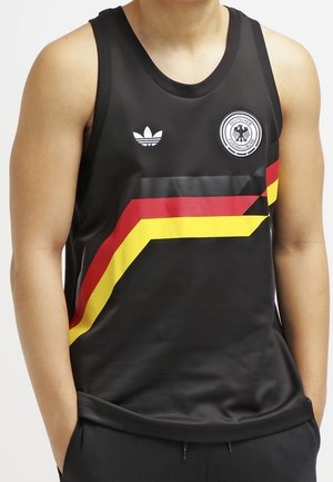 Mężczyzna ubrany w czarną sportową koszulkę na ramiączkach z diagonalnymi czerwonymi, żółtymi i czarnymi paskami oraz logo niemieckiej drużyny piłkarskiej na piersi.