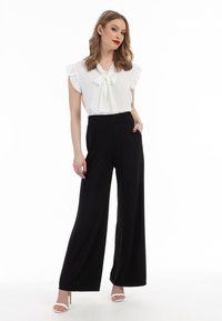 Weiße Bluse mit Rüschenärmeln und einem Schleifen-Ausschnitt, kombiniert mit hochtaillierten schwarzen weiten Hosen und weißen offenen High Heels.