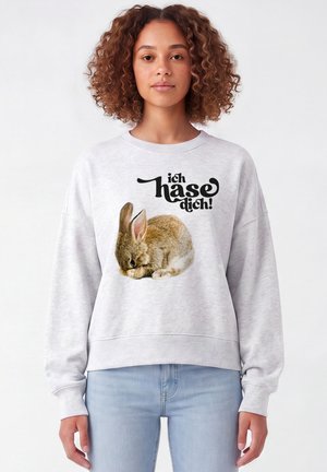 Jeune femme aux cheveux bouclés portant un sweat-shirt gris clair avec un lapin brun et un texte allemand "ich hase dich!" sur fond blanc.