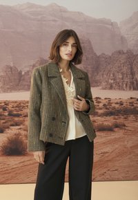 Brauner Blazer mit Fischgrätenmuster und doppelreihigen Knöpfen, aus leichtem Stoff, mit Reverskragen, kombiniert mit einer cremefarbenen Bluse und schwarzen Hosen.
