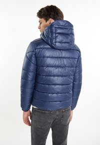 Námořnická modrá péřová bunda s kapucí, s prošívanou strukturou, lesklým povrchem a elastickými manžetami, nošená přes šedé džíny.