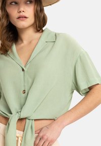 Camisa de manga corta verde claro con un frente atado, cierre de botones y tejido texturizado. Presenta un cuello con solapa y un corte relajado.