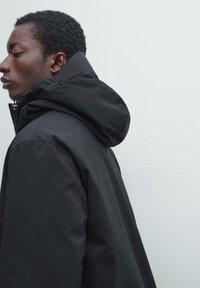 Czarna kurtka z kapturem o gładkiej, matowej fakturze; posiada wysoki kołnierz i zapięcie na zamek, widoczna z boku na jasnym tle.