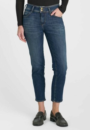 Femme portant un jean court bleu foncé avec fermeture à double bouton et mocassins noirs, debout avec une main dans la poche.