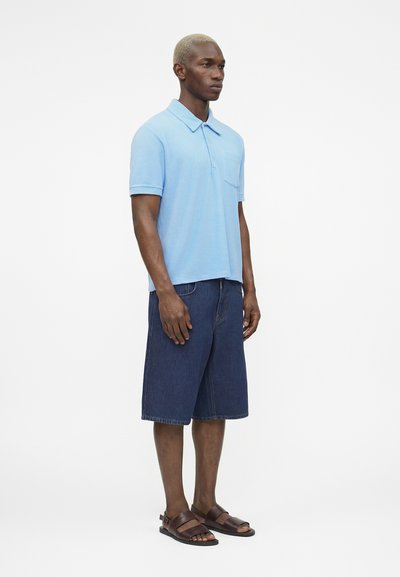 Άνδρας που φοράει ανοιχτό μπλε πόλο με κοντά μανίκια, σκούρα μπλε τζιν σορτς μέχρι το γόνατο και καφέ δερμάτινα σανδάλια, όρθιος πλάγια.