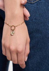 Bracciale in oro con un piccolo ciondolo rotondo e una pietra decorativa, indossato su una mano su uno sfondo di denim.