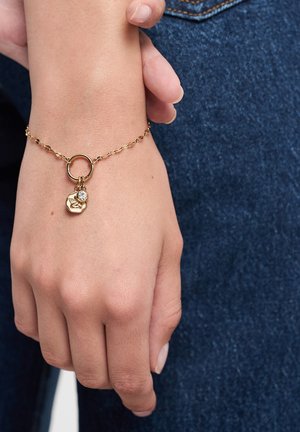 Gold chain bracelet featuring a small round charm with a stone accent, worn on a hand against a denim background.