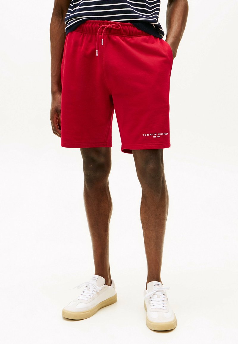 Homme portant un short rouge Tommy Hilfiger avec cordon, associé à des baskets blanches et une chemise rayée bleu marine et blanche.