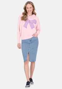 Sudadera rosa clara con un gráfico de un lazo morado, combinada con una falda lápiz de mezclilla con una abertura frontal. La modelo lleva zapatillas navy.