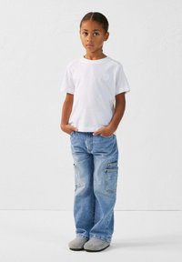 T-shirt blanche à manches courtes, jean cargo ample bleu clair avec poches latérales, et chaussures grises à enfiler. Design simple, style décontracté.