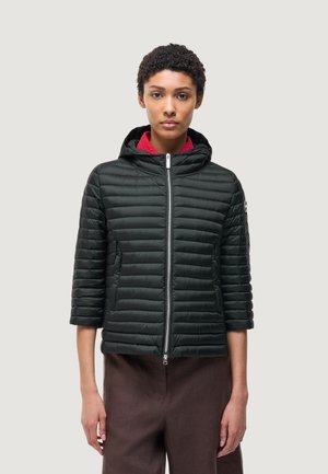 Donna con capelli corti e ricci che indossa una giacca trapuntata nera con cappuccio e maniche tre quarti, e pantaloni marroni, in piedi davanti a uno sfondo semplice.