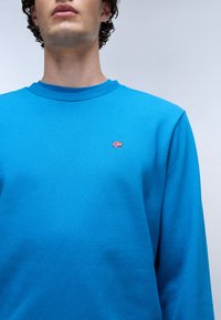 Felpa in cotone blu con scollatura a coste, caratterizzata da una piccola bandiera norvegese ricamata sul lato sinistro del petto. Texture liscia, maniche lunghe.