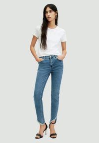 Jeans skinny azuis com bainhas desfiadas, combinados com uma t-shirt branca justa. O modelo está de pé com as mãos nos bolsos e a usar sandálias de salto pretas.