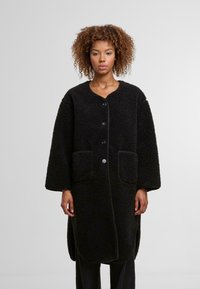 Donna con capelli ricci che indossa un lungo cappotto di pile nero con bottoni e tasche a toppa, in piedi contro uno sfondo grigio chiaro uniforme.