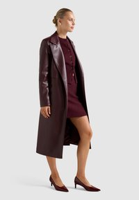 Μπουφάν από δέρμα βουργουνδί με γιακά, φορεμένο πάνω από ένα εφαρμοστό μίνι φόρεμα στο ίδιο χρώμα. Τα κλειστά τακούνια στο ίδιο χρώμα ολοκληρώνουν την εμφάνιση.