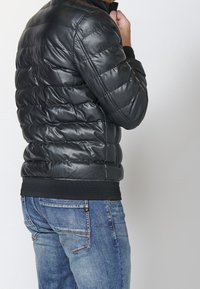 Homme en jean bleu portant une veste en cuir matelassée noire, levant une main près du menton sur un fond uni.