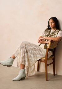 Crèmekleurige kanten rok met gestructureerde strepen, olijfgroene blouse met bloemmotief, en lichtblauwe enkellaarsjes met een gestapelde hak tegen een neutrale achtergrond.