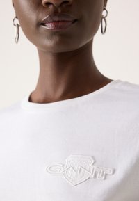 Gros plan d'une personne portant des boucles d'oreilles anneaux argentés et une chemise blanche avec un logo GANT blanc brodé sur la poitrine.
