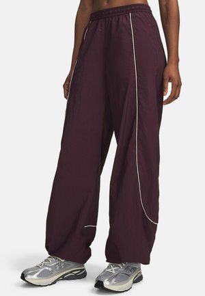 Person wearing loose, dark purple pants with white piping and silver athletic shoes, standing against a plain light background.