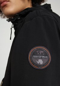 Schwarze Jacke mit Kapuze, aus strukturiertem Stoff, mit einem runden Aufnäher am linken Ärmel, der einen Globus und die Aufschrift "NAPAPIJRI" zeigt.