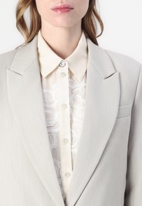 Beige skreddersydd blazer med skarpe slag, iført over en kremfarget skjorte med knapper og delikat broderidetalj. Sølvfarget knapp som aksent.