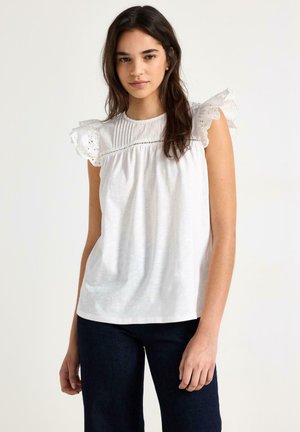 Ung kvinde med langt mørkt hår iført en hvid ærmeløs bluse med flæset blonder og mørke bukser, stående foran en ensfarvet baggrund.