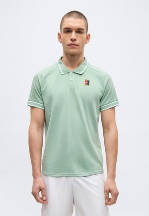 Joven de pie mirando al frente, lleva una camisa tipo polo verde claro de manga corta con logo en el pecho y pantalones cortos blancos sobre un fondo liso.