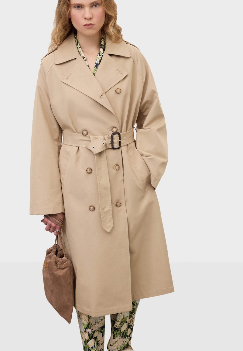 Femme portant un trench-coat beige ceinturé sur un pantalon floral, tenant un sac à cordon en daim marron dans sa main gauche.