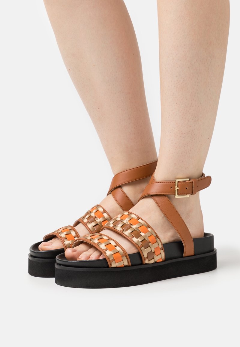 Stopy w noszących platformowych sandałach z czarnymi podeszwami, brązowymi paskami wokół kostki z złotymi klamrami oraz plecionych paskach z przodu w kolorach brązowym, pomarańczowym i złotym.