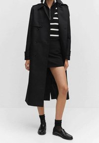 Czarny płaszcz typu trencz z podwójnym zapięciem, wykonany z lekkiego materiału, z pagonami na ramionach, noszony na czarno-białym swetrze w paski i mini spódniczce.