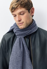 Dunkelgrauer Fischgrätenmuster-Schal, locker um den Hals drapiert, getragen über einer schwarzen, strukturierten Lederjacke, die ihr weiches Material betont.