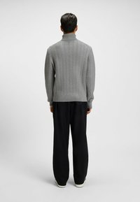 Grauer Strickpullover mit hohem Kragen, der ein Fischgrätmuster aufweist. Kombiniert mit lockeren schwarzen Hosen und hellen Schuhen.