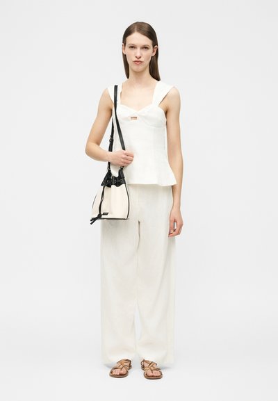 Jonge vrouw met een witte mouwloze top, wijde witte broek, bruine sandalen, en een zwart-witte bucketbag over de schouder.