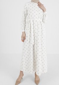 Robe blanche à manches longues avec des pois marron, boutonnière sur le devant, ceinture à la taille cintrée et jupe fluide avec une coupe ample.