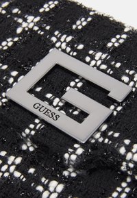 Tissu en tweed noir et blanc présentant un logo en métal "G" bien visible. Surface texturée avec des brins et un motif tissé sur fond noir.