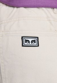 Pantalons en coton beige avec une étiquette noir et blanc représentant deux mains dans un design rectangulaire sur la poche arrière.