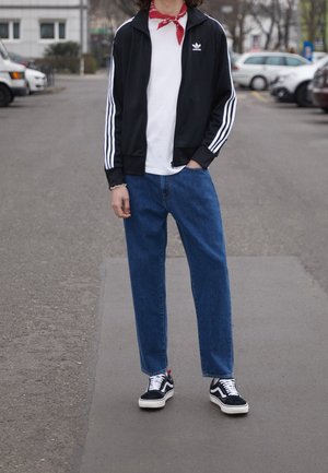 Persona in piedi per strada con giacca Adidas nera, maglietta bianca, jeans blu, scarpe da ginnastica nere e sciarpa rossa al collo.