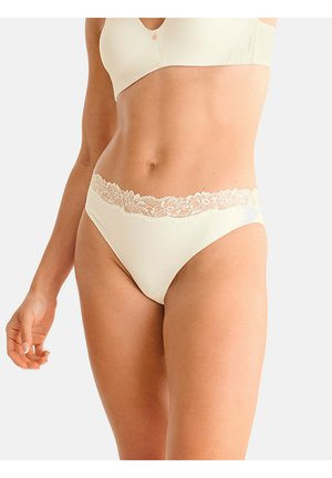 Femme portant un bikini crème orné de dentelle et un soutien-gorge doux assorti, debout devant un fond clair uni.