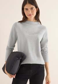 Hellgrauer Pullover-Sweatshirt mit langen Ärmeln, das ein geprägtes Textmuster aufweist. Schwarze leichte Pufferjacke unter dem Arm gehalten.