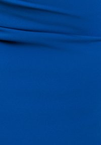 Sininen kangas, jossa on pehmeitä taitoksia ja sileä pinta, tasaisesti valaistu, näyttäen hienovaraisia varjoja ja korosteita.