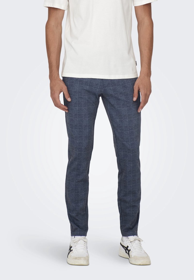 Pantalon à motifs marine avec un design à carreaux, coupe slim et texture douce, associé à un t-shirt neutre et des baskets blanches.