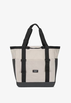 Taška přes rameno s béžovou horní částí, černým dnem a strapci. Vyrobena z odolné látky s hladkou texturou a logem na přední straně.