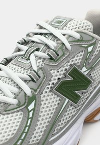 Chaussure de sport grise en matériau mesh avec des accents verts. Présente des lacets blancs, un logo vert bien en vue et une semelle rembourrée.
