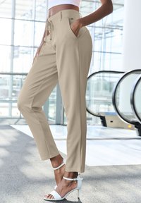 Beige op maat gemaakte broeken van gladde stof, met zijzakken en een tailleband met trekkoord, gecombineerd met witte hoge hakken sandalen.