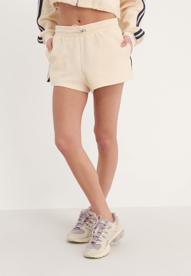 Shorts beige con textura acanalada, cintura elástica y bolsillos laterales, combinados con zapatillas deportivas grises y moradas. La tela tiene un acabado texturizado.