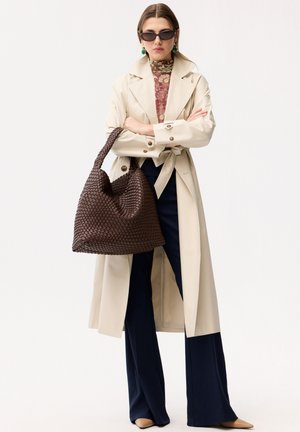 Donna con trench beige, top con motivo, pantaloni blu navy, che tiene una grande borsa marrone intrecciata, indossa occhiali da sole scuri e orecchini verdi.